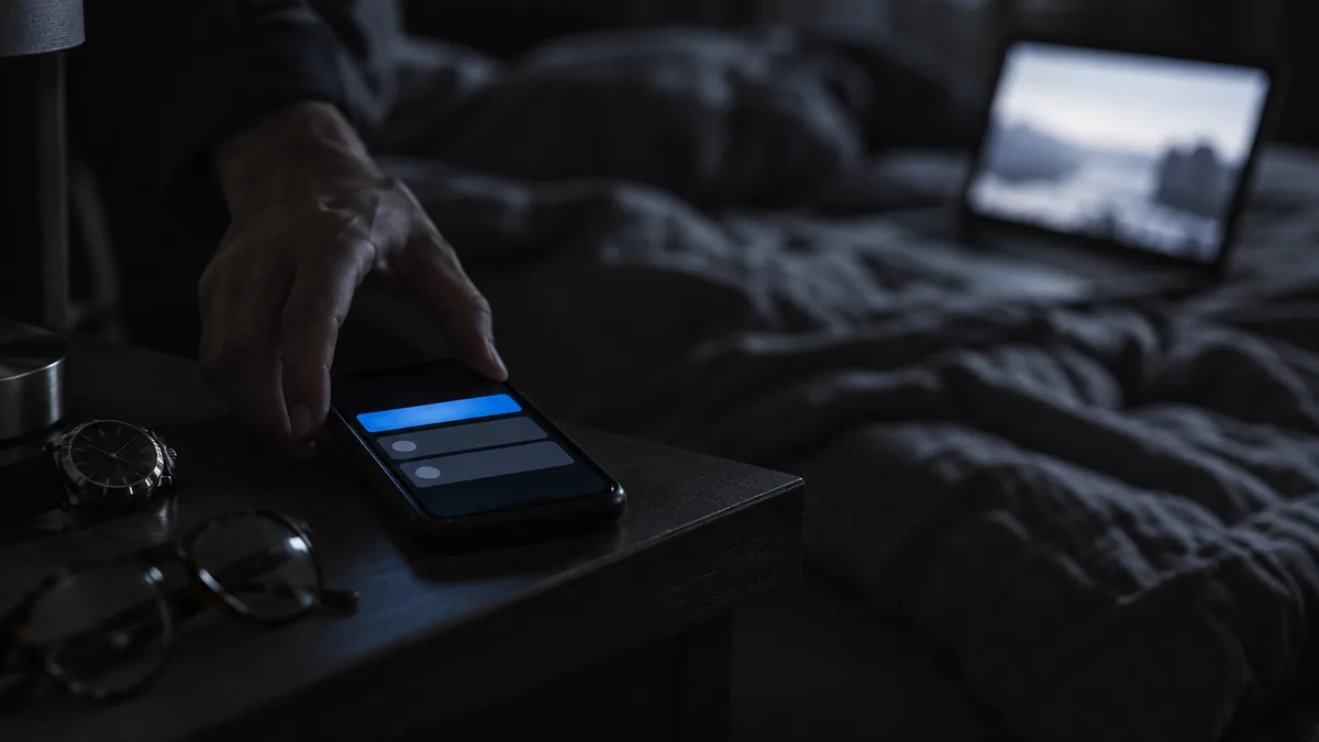 Téléphone d'astreinte allumé et vibrant sur une table de chevet la nuit, à côté d'une montre et de lunettes, une main s'en saisissant, un ordinateur ouvert sur le lit au fond.